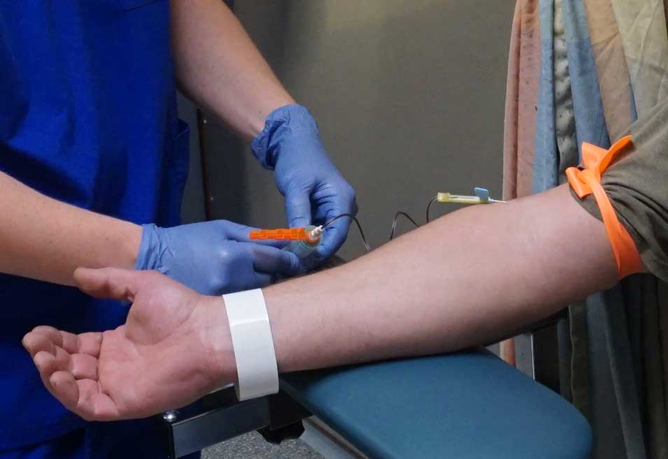 Healthcare professional in blue scrubs preparing to draw blood from a patient's arm using a tourniquet.
