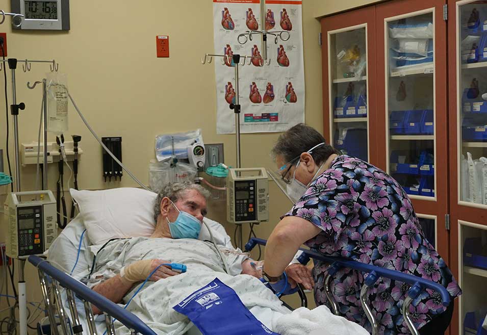 Healthcare professional wearing scrubs, face shield, and mask tending to a patient lying in a hospital bed in an emergency room setting.