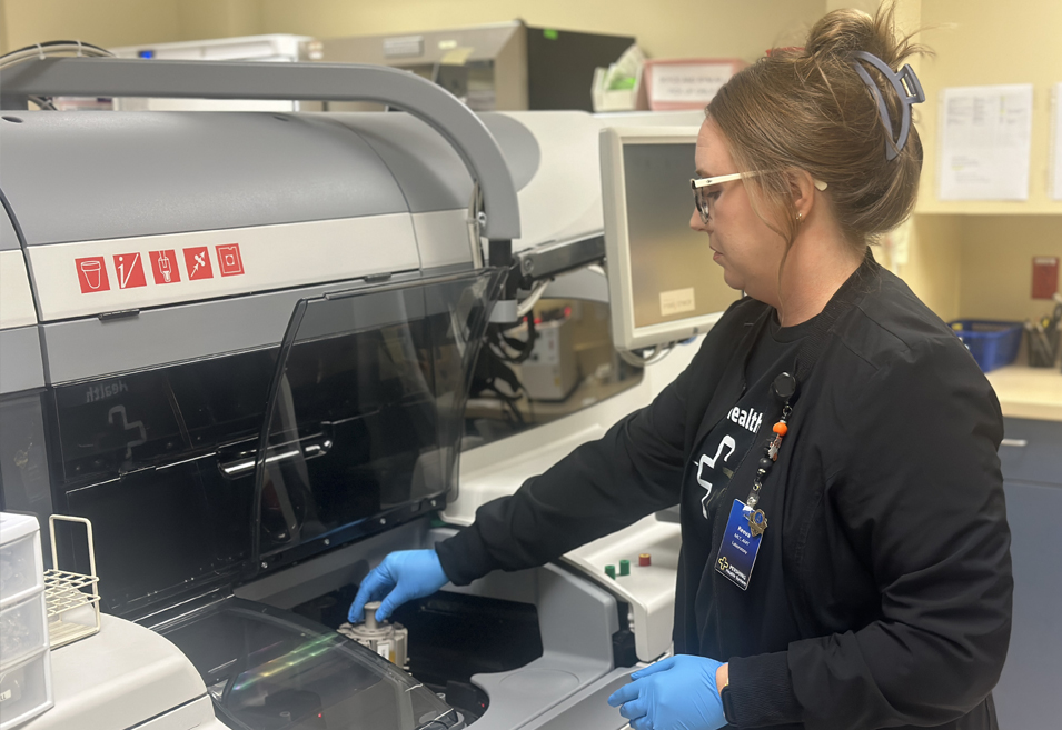 Reeva wearing safety glasses and blue gloves while operating a laboratory analyzer machine.