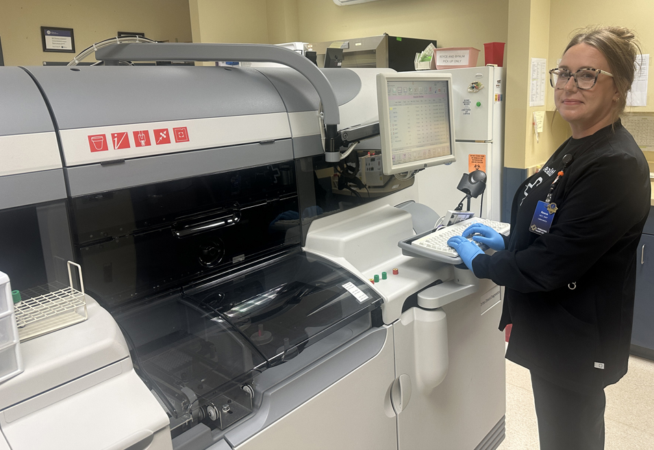 Reeva standing next to a laboratory analyzer machine in a clinical testing facility.