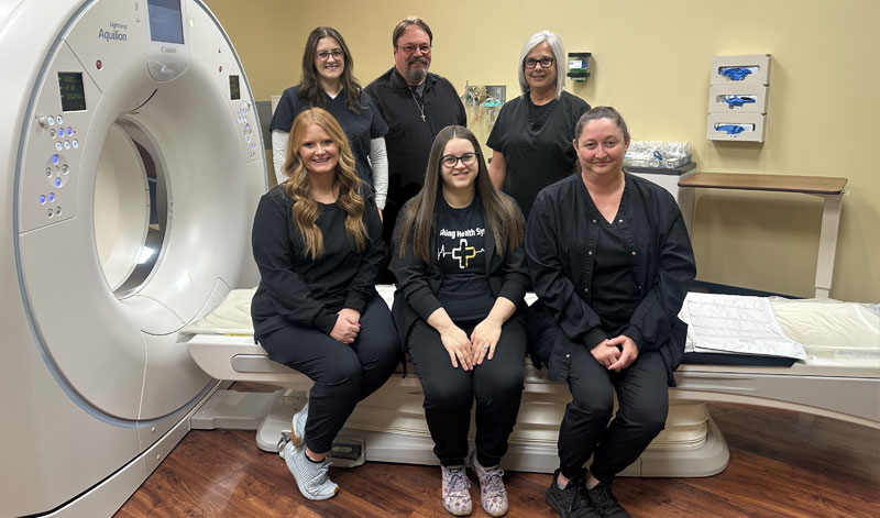 Six staff members posing together in front of a CT scanner in a medical imaging room.