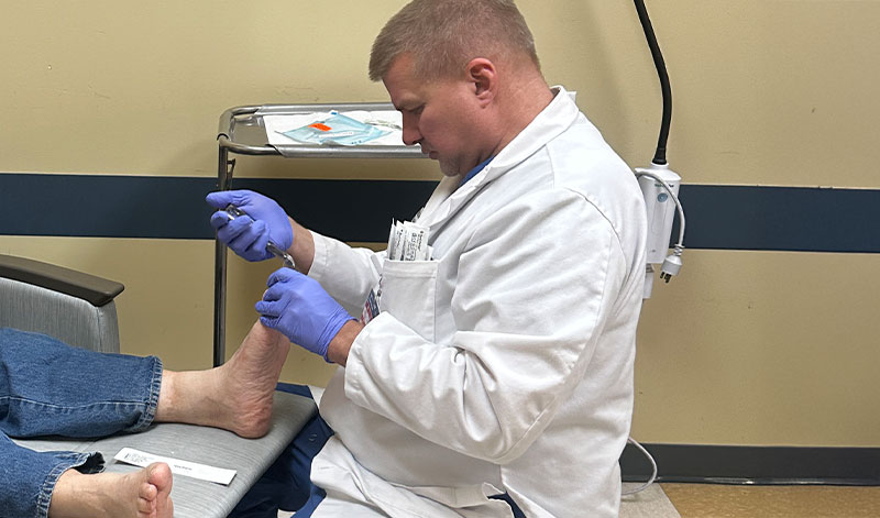 Healthcare professional wearing a white coat and blue gloves examining a patient's foot in a clinical setting.