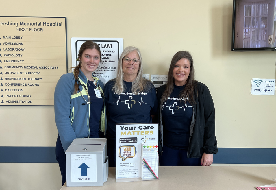 Three staff members standing behind a table displaying a patient comment box and a sign that reads, Your Care Matters Tell Us!